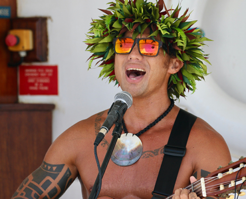 Sänger mit einer Pflanzenkrone, Sonnenbrille und Tattoos, er spielt dabei Gitarre